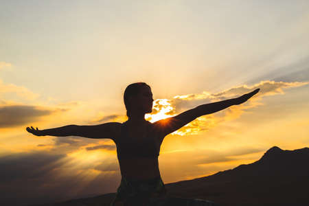 Silhouette of young woman practicing yoga or pilates at sunset or sunrise in beautiful mountain location, doing lunge exercise, standing in Warriorの写真素材