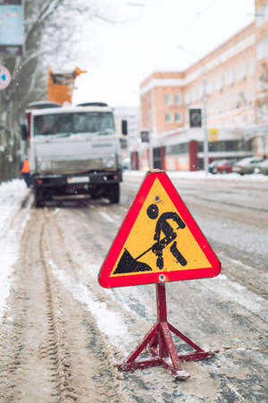 Snow-plow remove snow from the city street.Warning road sign.Winter service vehicle snow blower work.Cleaning snowy frozen roads.の写真素材