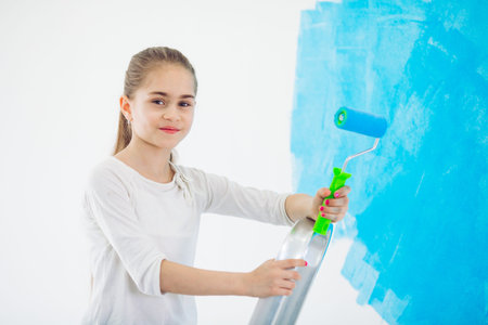Repair in the apartment. Happy child girl paints the wall with blue paint,