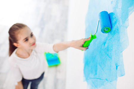 Repair in the apartment. Happy child girl paints the wall with blue paint,
