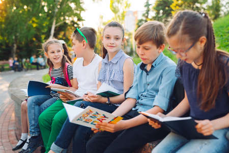 Happy Schoolmates Portrait. Schoolmates seating with books in a wooden bench in a city park and studying on sunny day.の写真素材