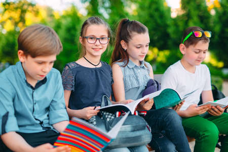 Happy Schoolmates Portrait. Schoolmates seating with books in a wooden bench in a city park and studying on sunny day.の写真素材