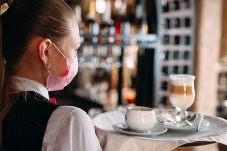 A female Waiter of European appearance in a medical mask serves Latte coffee.