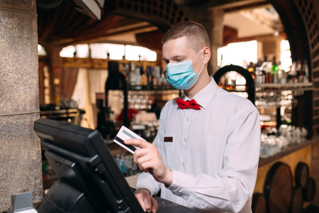 small business, people and service concept. man or waiter in medical mask at counter with cashbox...