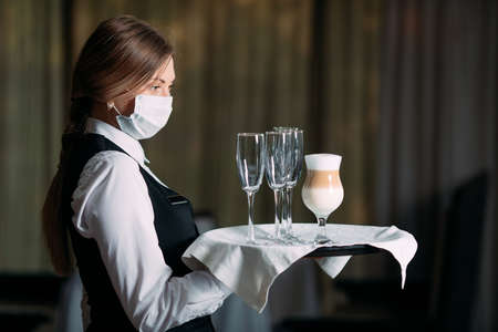 A female Waiter of European appearance in a medical mask serves Latte coffee.の写真素材