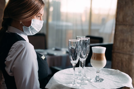 A female Waiter of European appearance in a medical mask serves Latte coffee.