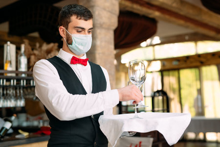 A waiter in a medical protective mask serves the table in the restaurant.