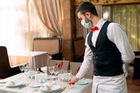 A waiter in a medical protective mask serves the table in the restaurant.