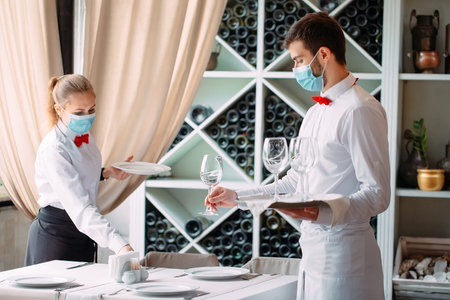 A waiter in a medical protective mask serves a table in the restaurant. Employees of a restaurant or...