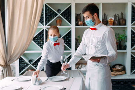 A waiter in a medical protective mask serves a table in the restaurant. Employees of a restaurant or hotel in protective masks. The End Of Quarantine.の写真素材