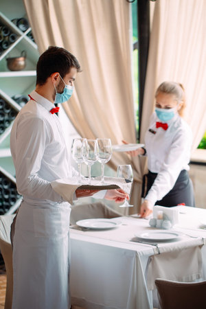 A waiter in a medical protective mask serves a table in the restaurant. Employees of a restaurant or...