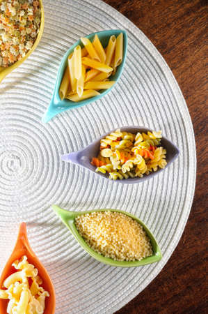 various types of uncooked pasta in ceramic containers on a white tablecloth top viewの写真素材