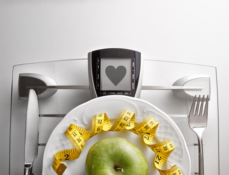 Cutlery with yellow tape measure and green apple on a scale plate on a white table. Concept of healthy eating at the table with healthy heart message. Horizontal composition. Top view.の写真素材