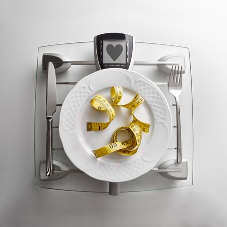 Cutlery with yellow tape measure on a scale plate on a white table. Concept of healthy eating at the table with healthy heart message. Square composition. Top view.の写真素材