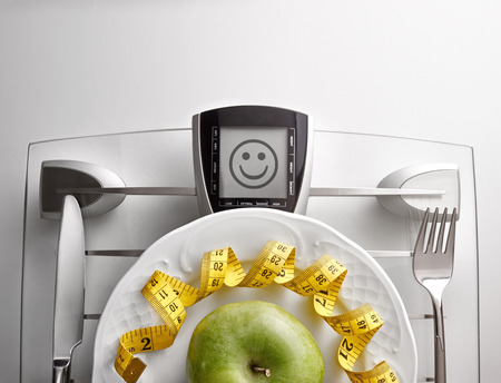 Cutlery with yellow tape measure and green apple on a scale plate on a white table. Concept of healthy eating at the table with happy face message. Horizontal composition. Top view.の写真素材