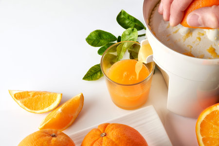 Hand squeezing an orange with an electric juicer in a glass on a white kitchen table. Oranges on a...