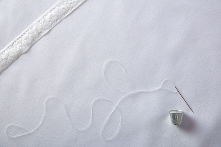 Background sewing needle threaded with white thread thimble and lace ribbon on white cloth. Horizontal composition. Top view.の写真素材