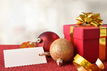 Xmas decoration with tag, red gift box and golden ribbon, and two balls on bright red textured table and white background. Front view.の写真素材