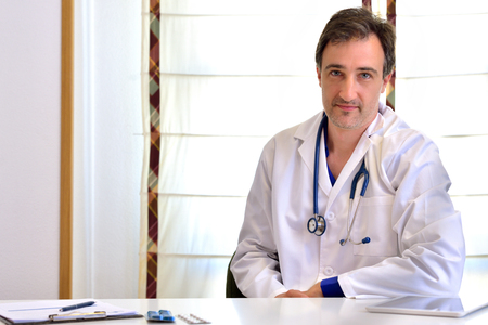 Caucasian general doctor in consultation front position prescribe medication. White background with blind and tablet, clipboard and  on white table. Horizontal composition.の写真素材