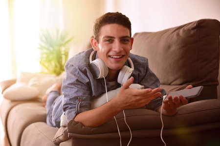 Teen listening music with headphones from mobile lying face down on couch surprised and laughing