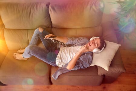 Teenager listening to music with headsets from a smartphone and dancing face up on couch in the living room with multicolored lights. Top viewの写真素材