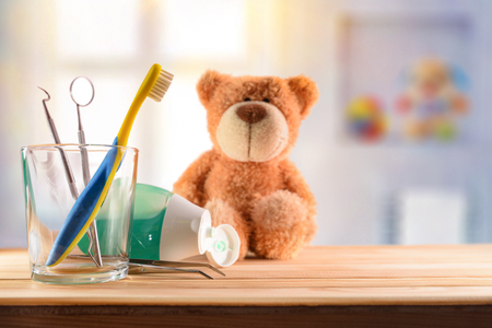 Dentist concept for children with tools in glass cup and teddy on wooden table in room with toys background. Horizontal composition. Front viewの写真素材