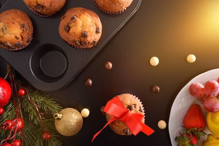 Freshly homemade baked chocolate muffins on mold on black table with christmas decoration and a dish of fruit. Horizontal composition. Top view.の写真素材