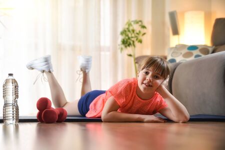 Smiling girl resting on a mat on the floor looking at a camera happy in a sport session in the living room at home.の写真素材