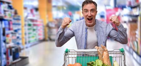 Happy supermarket customer raising hands with clenched fists with gesture of success with shopping cart full of food in supermarket aisleの写真素材