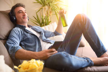 Relaxed man listening to music with headphones from a mobile phone with a beer in his hand sitting on a sofa at home comfortablyの写真素材