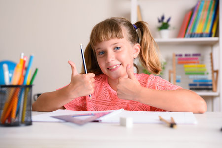 Smiling girl studying at home with raised thumbs with desk with educational material.