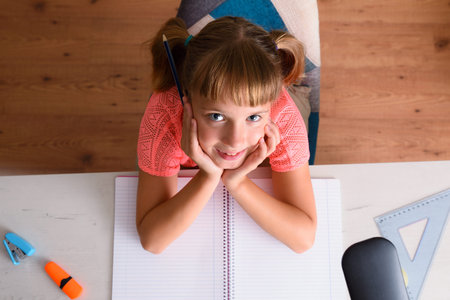 Smiling little girl studying with her hands on chin on desk. Top view.の写真素材
