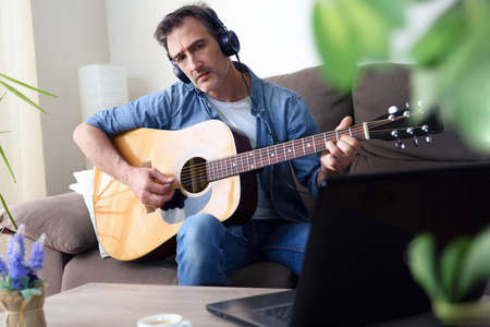 Man listening to music with headphones and playing the guitar looking at a laptop at the same time in the living room at home detailの写真素材