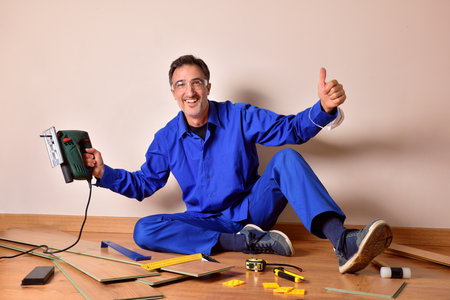 Professional parquet assembler with ok gesture and cutting machine in hand sitting on the floor assembling parquet. Horizontal composition.の写真素材