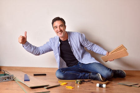 Man smiling with hand with ok gesture sitting on the floor putting parquet in his own house. Horizontal composition.の写真素材