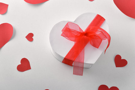 White gift box with heart shape and red bow and cutouts of red hearts on white table. Elevated view.の写真素材