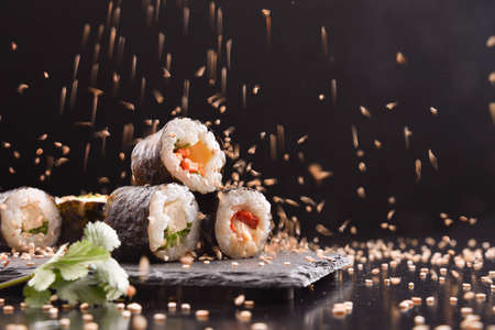 Detail of assorted sushi maki on slate plate and chopsticks on black wooden table and sesame seeds falling on top. Front view. Horizontal composition.の写真素材