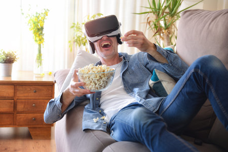 Man watching multimedia content in virtual reality glasses eating popcorn lying on the sofa at home