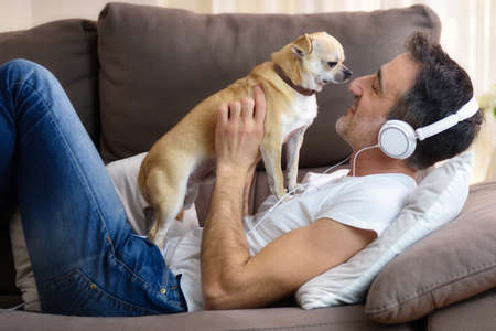 Man playing with his dog affectionately looking at his face lying on the sofa at home listening to music with white headphones close up.の写真素材