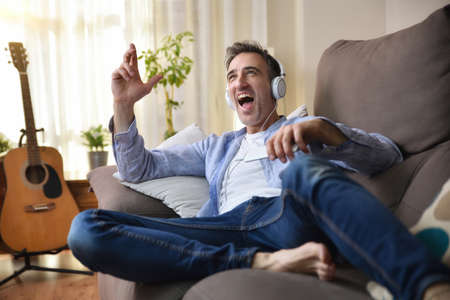 Euphoric adult man laying dancing in sofa listening to music from a mobile with white headphones dressed casual in the living room with window and background guitarの写真素材