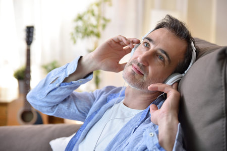 Attentive man listening through headphones sitting on his sofa at home with a window in the...