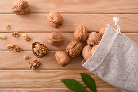 Group of walnuts spilling out of a cloth sack and seeds and leaves on wooden table. Top view.