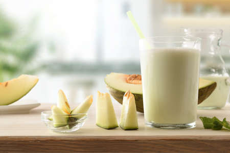 Glass with natural melon milk shake on a plate on wooden kitchen bench with cut fruit. Front view.の写真素材