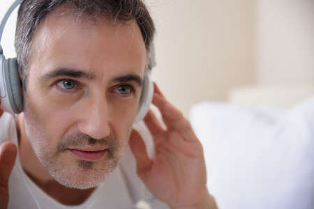 Closeup of the head of a concentrated man listening through white headphones with gray pads in a roomの写真素材