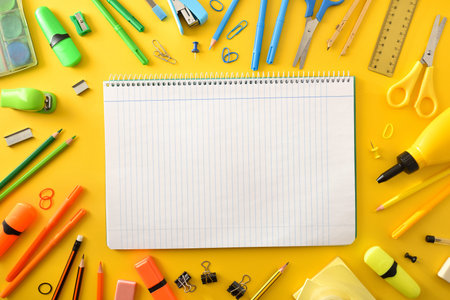 Open school line notebook in the center with school supplies around on yellow background. Top view.