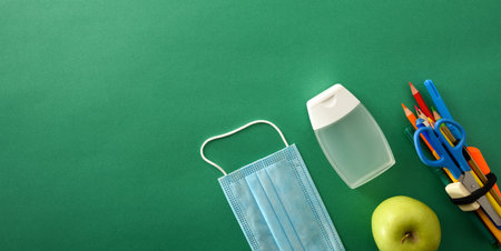 School tools with apple with mask and hydroalcoholic gel on green background with space for text. Top view. Panoramic composition.の写真素材