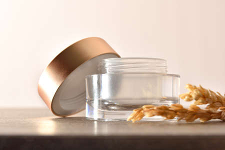 Open glass jar for empty face or body cream with golden lid on wooden table and oat spikes with beige isolated backgroundの写真素材