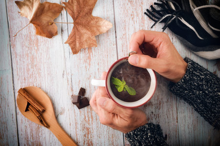 Cold hands with wool sweater stirring hot chocolate in a mug on wooden table with chocolate and...