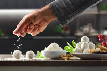 Hand pouring grated coconut over home prepared coconut balls on kitchen bench with ingredients and...