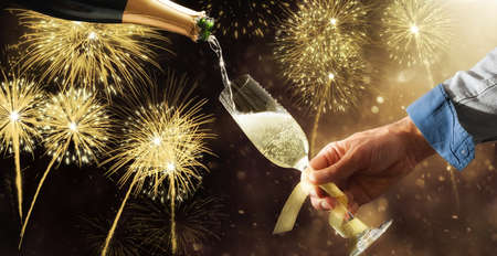 Filling champagne glass adorned with gold bow from a bottle with fireworks background at a party. Front view.の写真素材